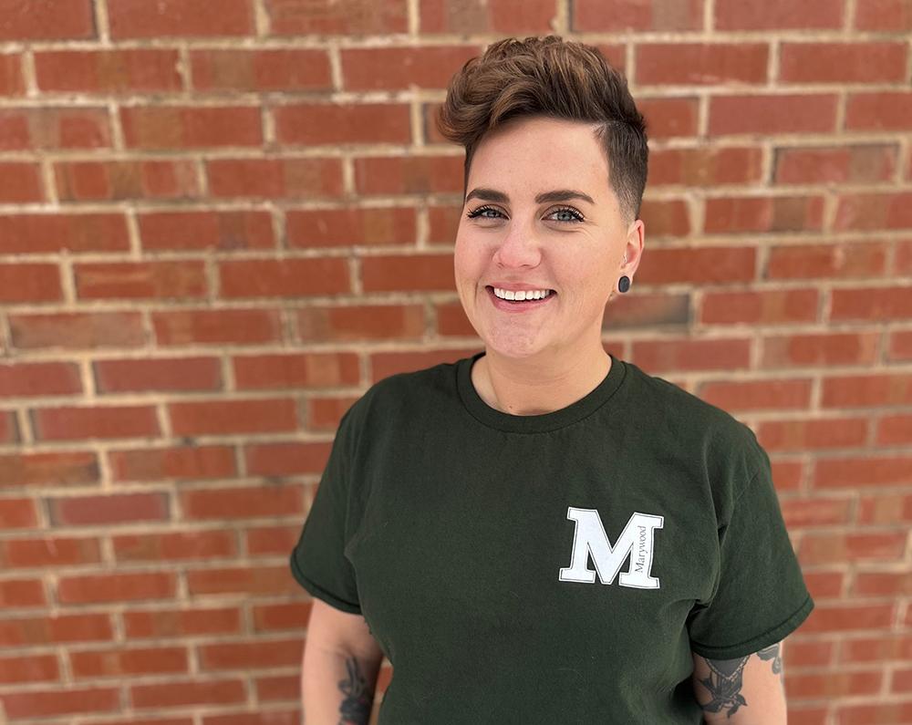 Woman smiling wearing. green marywood t-shirt with a brick wall in the background.